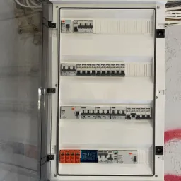 Otwarta skrzynka rozdzielcza z zamontowanymi wyłącznikami nadprądowymi i zabezpieczeniami przepięciowymi na ścianie z widocznymi kablami i śladami farby.