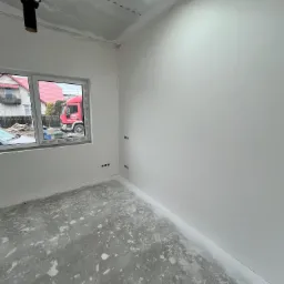 Wnętrze pokoju po szpachlowaniu ścian i sufitu, widoczne gładkie, białe powierzchnie, surowa podłoga z resztkami zaprawy i instalacja wentylacyjna pod sufitem. Przez okno widoczny czerwony samochód...