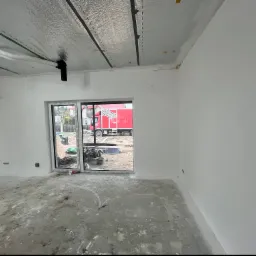 Wnętrze w trakcie wykończenia: ocieplone poddasze z widoczną folią aluminiową, świeżo pomalowane ściany na biało, betonowa wylewka na podłodze, okno z widokiem na czerwony samochód dostawczy.