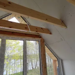 Poddasze w trakcie wykańczania: widoczne drewniane belki konstrukcyjne, płyty kartonowo-gipsowe na skosach oraz okna z widokiem na zieleń i kwitnące żółte kwiaty na zewnątrz.