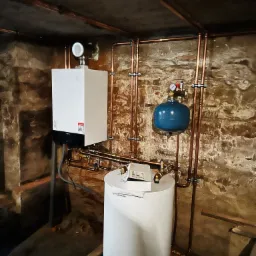 Nowoczesny piec gazowy Viessmann, podłączony miedzianymi rurami, z niebieskim zbiornikiem wyrównawczym, zamontowany w piwnicy o kamiennych ścianach.