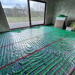 Instalacja ogrzewania podłogowego z zielonych rurek na czarnej folii z czerwoną siatką, widok z góry na surowy pokój z dużym oknem i widokiem na drzewa.