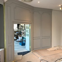 ATG InterWorks - Wnętrze w trakcie remontu: szare ściany z dekoracyjnymi panelami, zabezpieczone taśmą malarską, widoczne kable na podłodze i otwarte drzwi do zagraconego pomieszczenia.