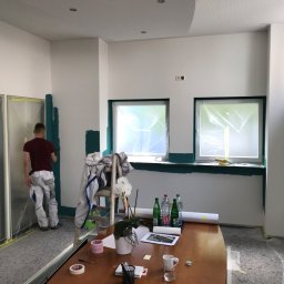ATG InterWorks - Mężczyzna w roboczym ubraniu maluje na zielono fragment ściany w biurze, zabezpieczone okna folią, na stole butelki wody, dokumenty i przybory biurowe.