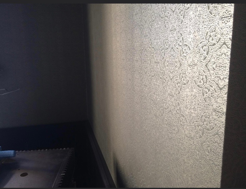 Fragment ściany w rogu pokoju pokrytej jasną tapetą w orientalne wzory, z widoczną listwą przypodłogową i fragmentem podłogi.