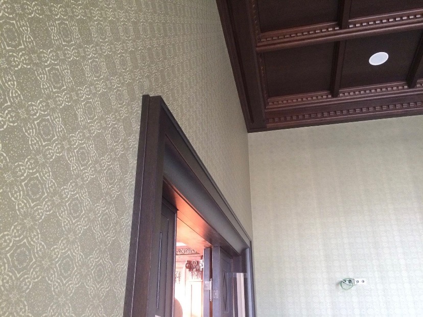 Fragment wnętrza: ściana pokryta tapetą w delikatny wzór, ciemna, drewniana rama drzwi, dekoracyjny sufit kasetonowy z oświetleniem punktowym.