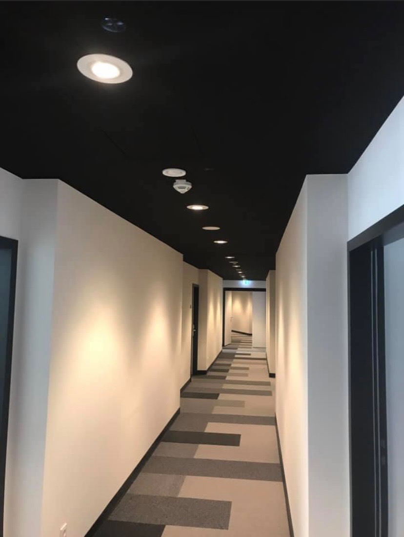 Korytarz biurowy w Warszawie z białymi ścianami, czarnym sufitem i wpuszczanymi lampami LED, wykończony wykładziną dywanową w geometryczne wzory.