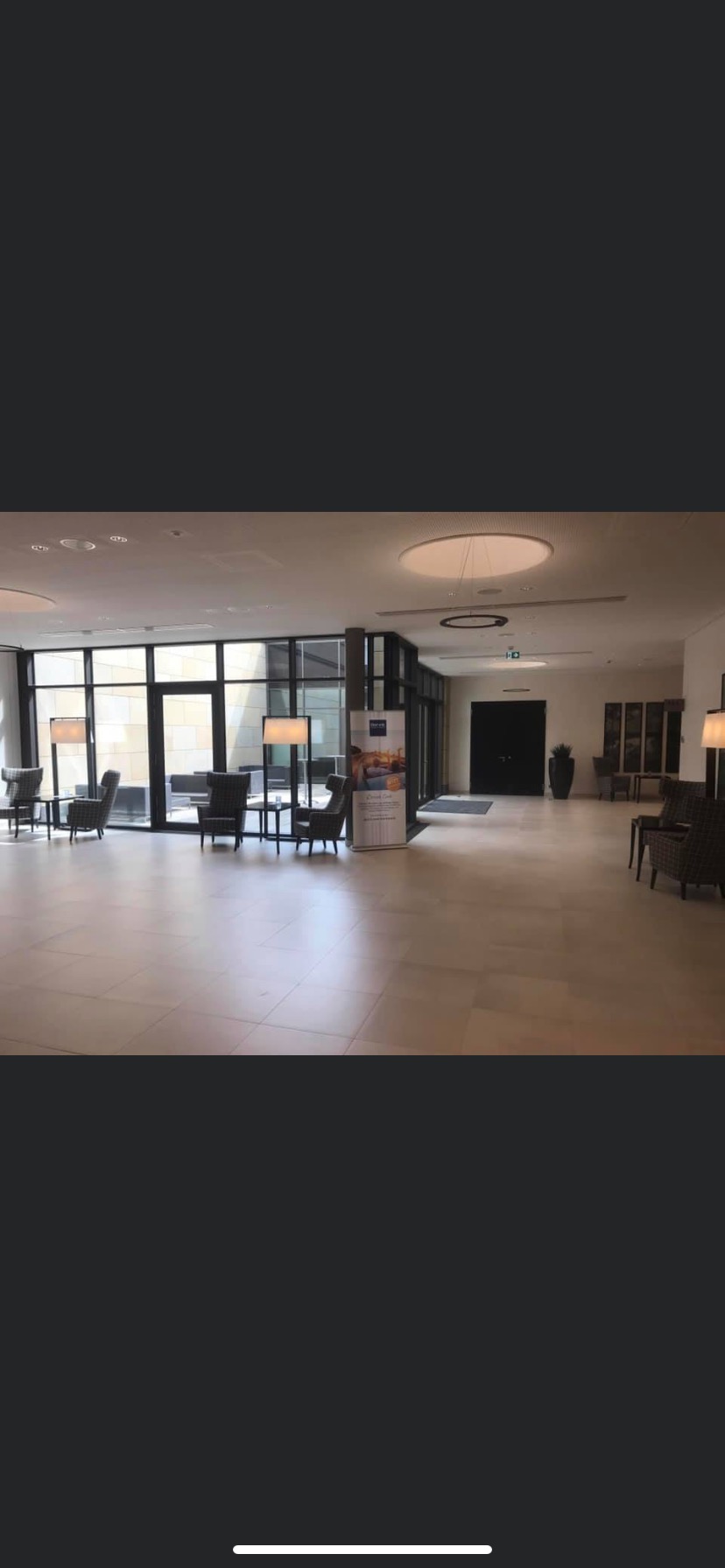 Jasne, przestronne lobby biurowe z dużymi oknami, nowoczesnymi lampami stojącymi i komfortowymi fotelami, z widocznym plakatem reklamowym w tle.