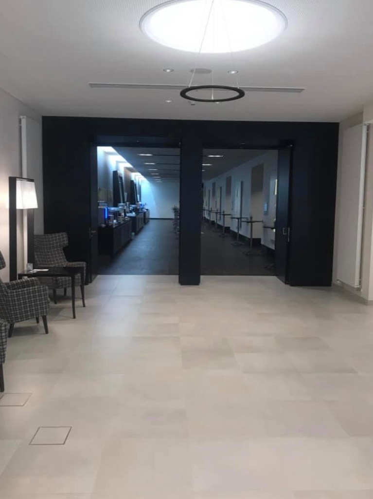 Wnętrze biurowe z jasną podłogą, czarnymi framugami drzwi i nowoczesnym oświetleniem sufitowym, widok na korytarze z wyposażeniem.