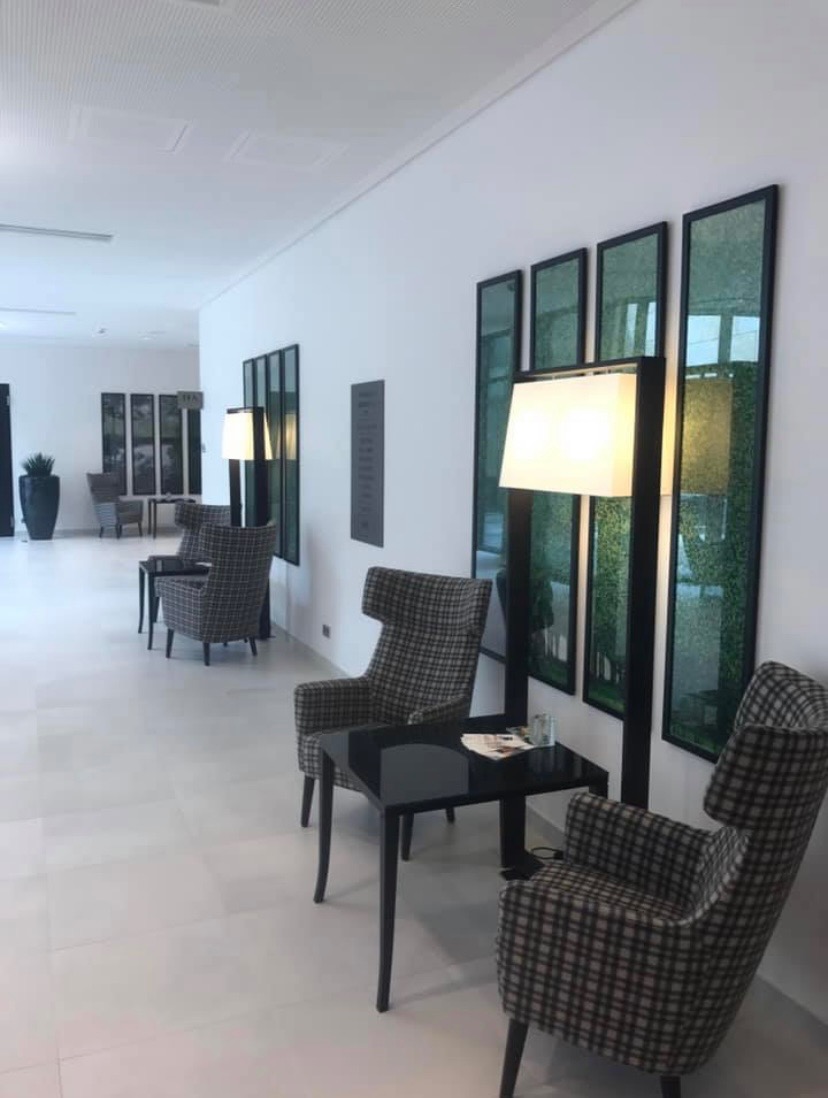 Minimalistyczne wnętrze biurowe z białymi ścianami, szarymi panelami dekoracyjnymi, szarymi fotelami w kratę i czarnymi stolikami kawowymi, oświetlone lampami podłogowymi.