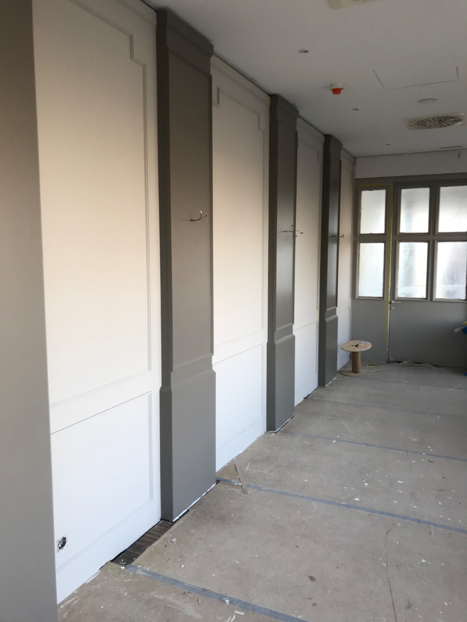 Korytarz z panelami ściennymi w kolorze białym i szarym, zakończone malowaniem, widoczne haki na ścianie i przygotowane podłoże.