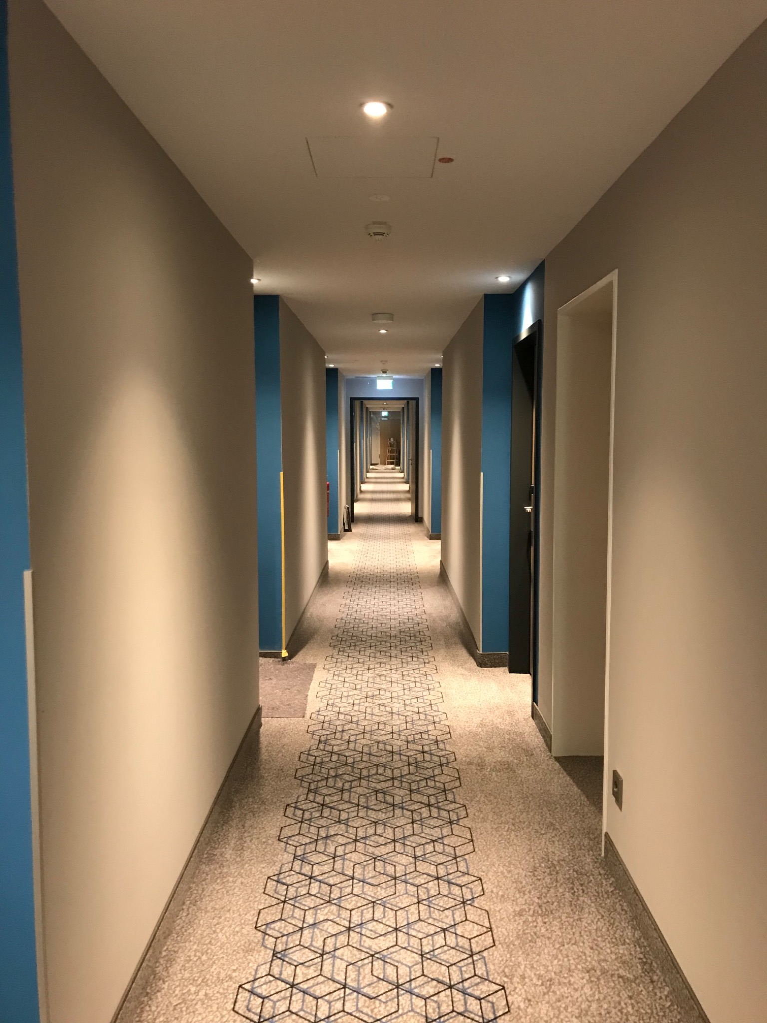 Długi korytarz biurowy z beżowymi ścianami, niebieskimi akcentami i wzorzystą wykładziną podłogową w geometryczne sześciany, oświetlony punktowymi lampami sufitowymi.
