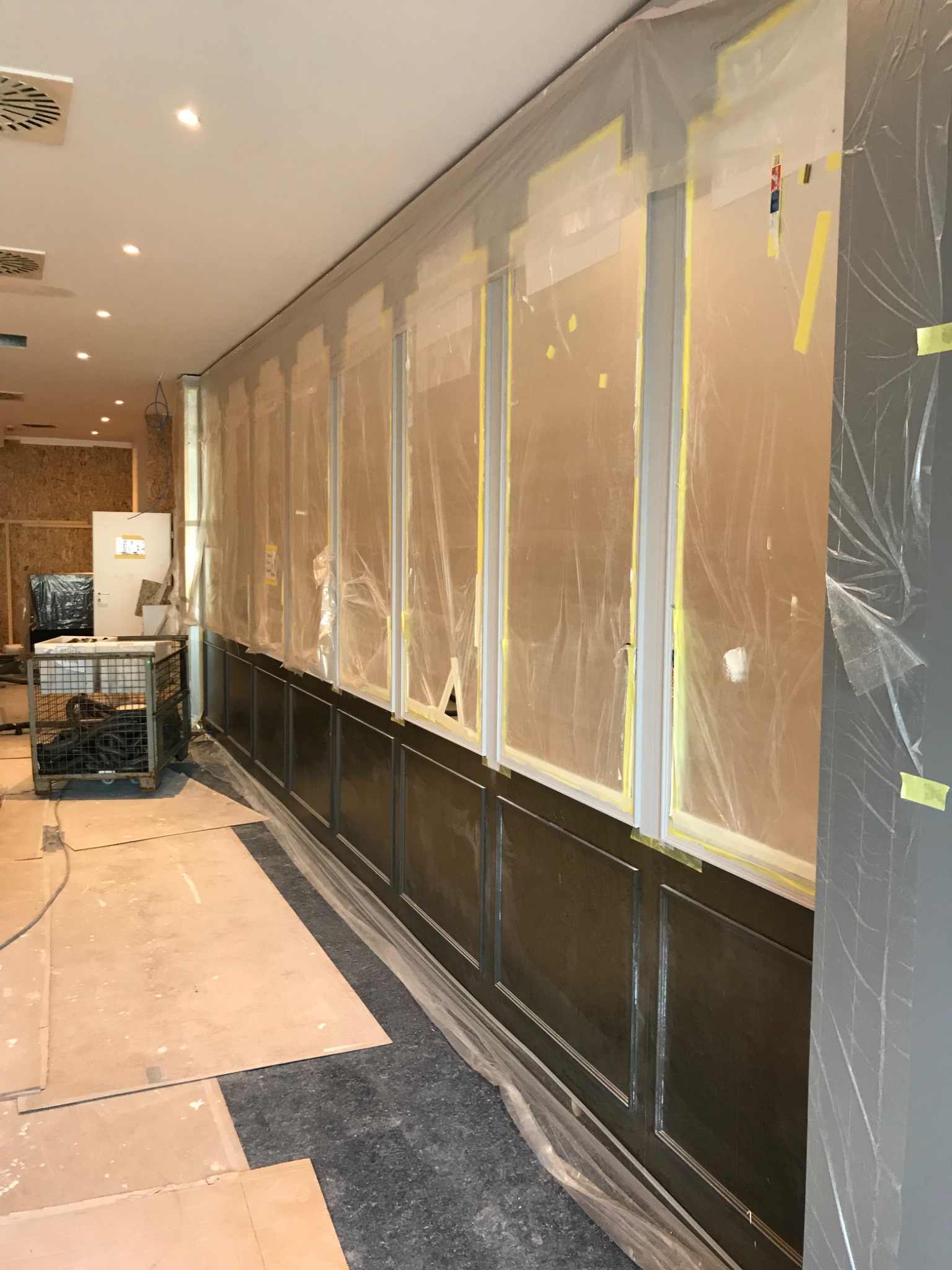 Przygotowanie do odświeżenia ścian w lokalu: dolna część ściany w ciemnym drewnie, górna część zabezpieczona folią malarską i taśmą, widoczne kartony na podłodze i metalowy wózek z przewodami.