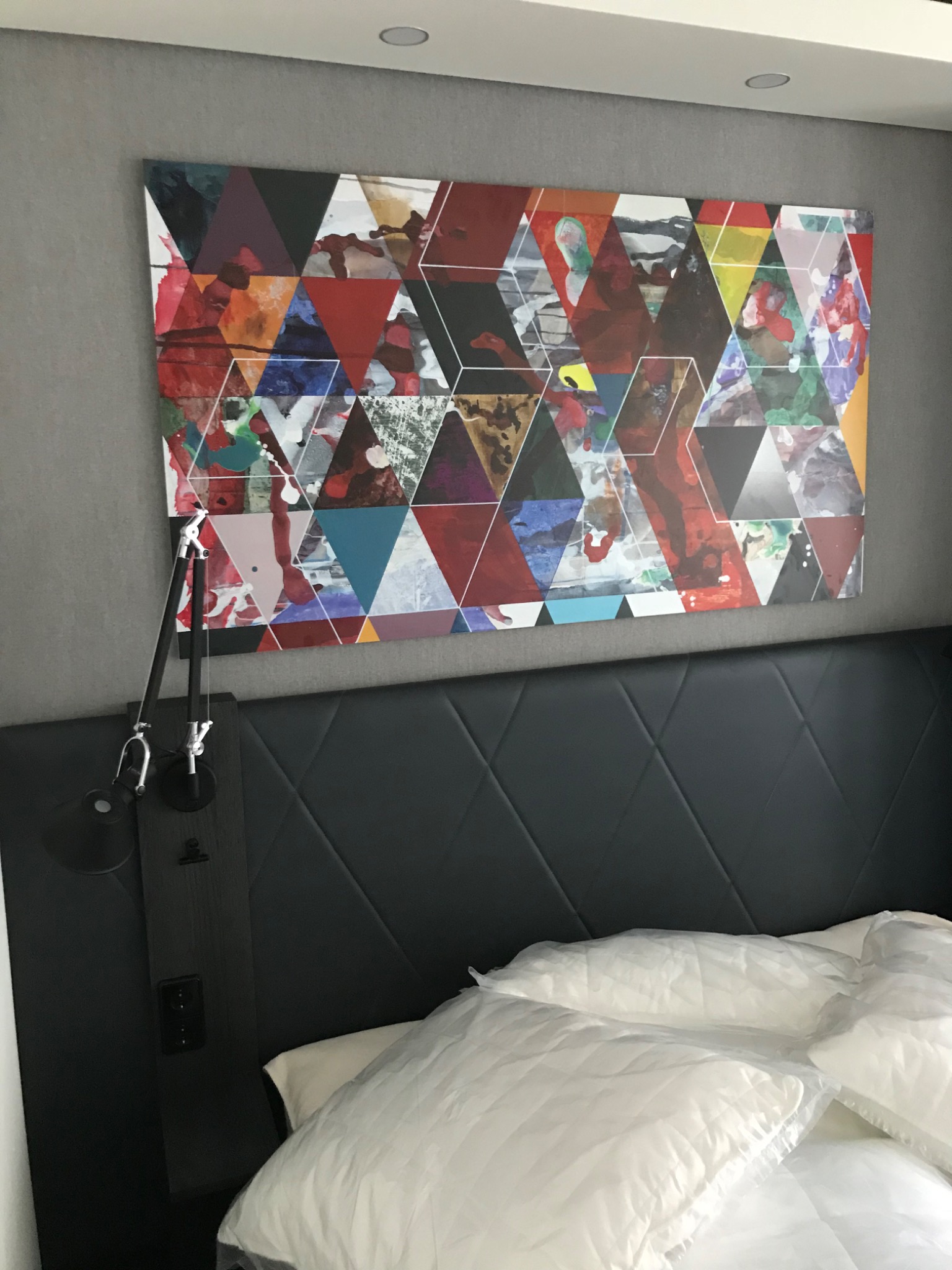 Abstrakcyjny obraz geometryczny z trójkątami w kolorach czerwonym, niebieskim, zielonym i żółtym, zawieszony nad wezgłowiem łóżka w sypialni, oświetlony lampką nocną