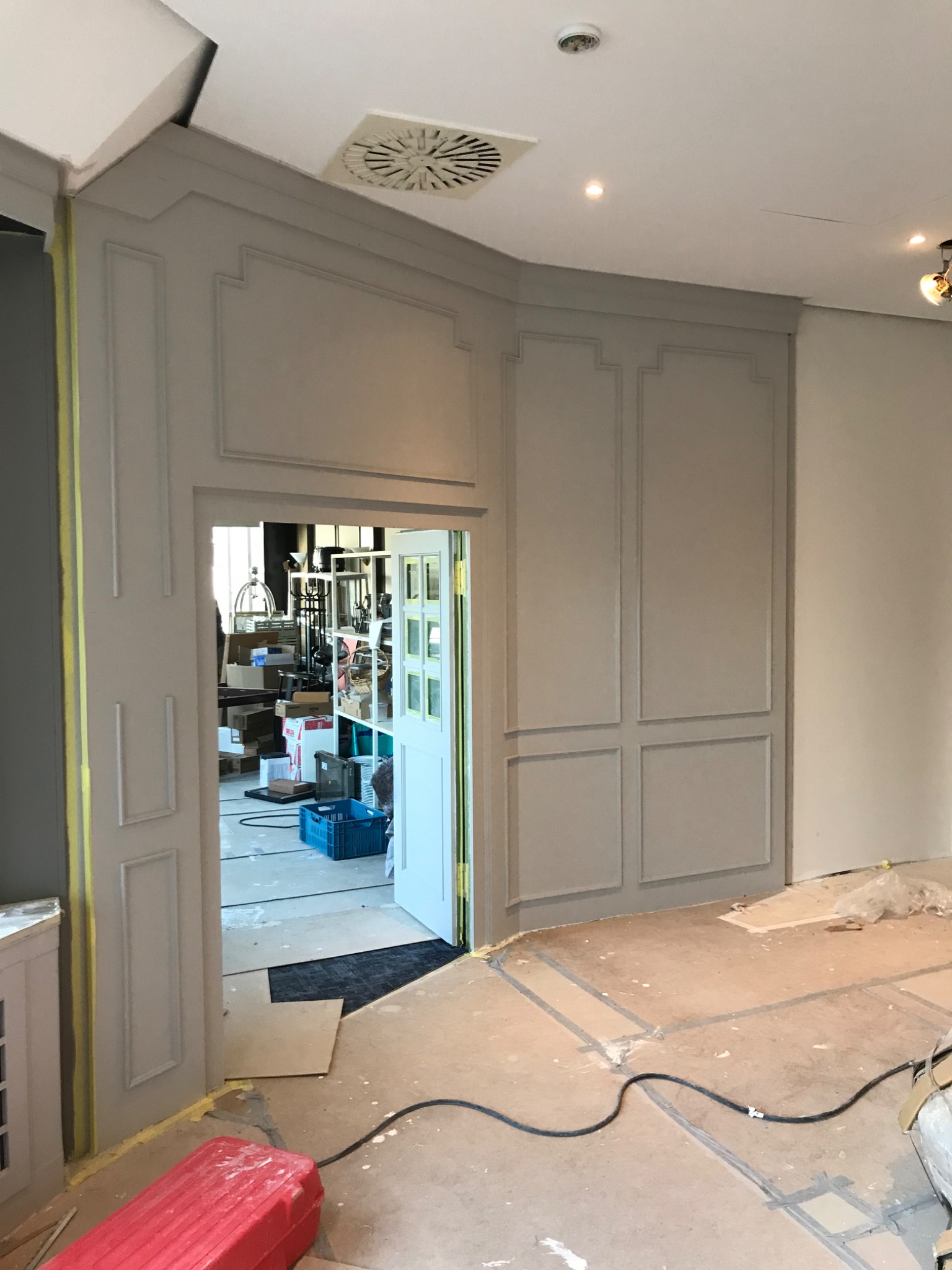 Wnętrze w trakcie remontu: szare ściany z dekoracyjnymi panelami, zabezpieczone taśmą malarską, widoczne kable na podłodze i otwarte drzwi do zagraconego pomieszczenia.