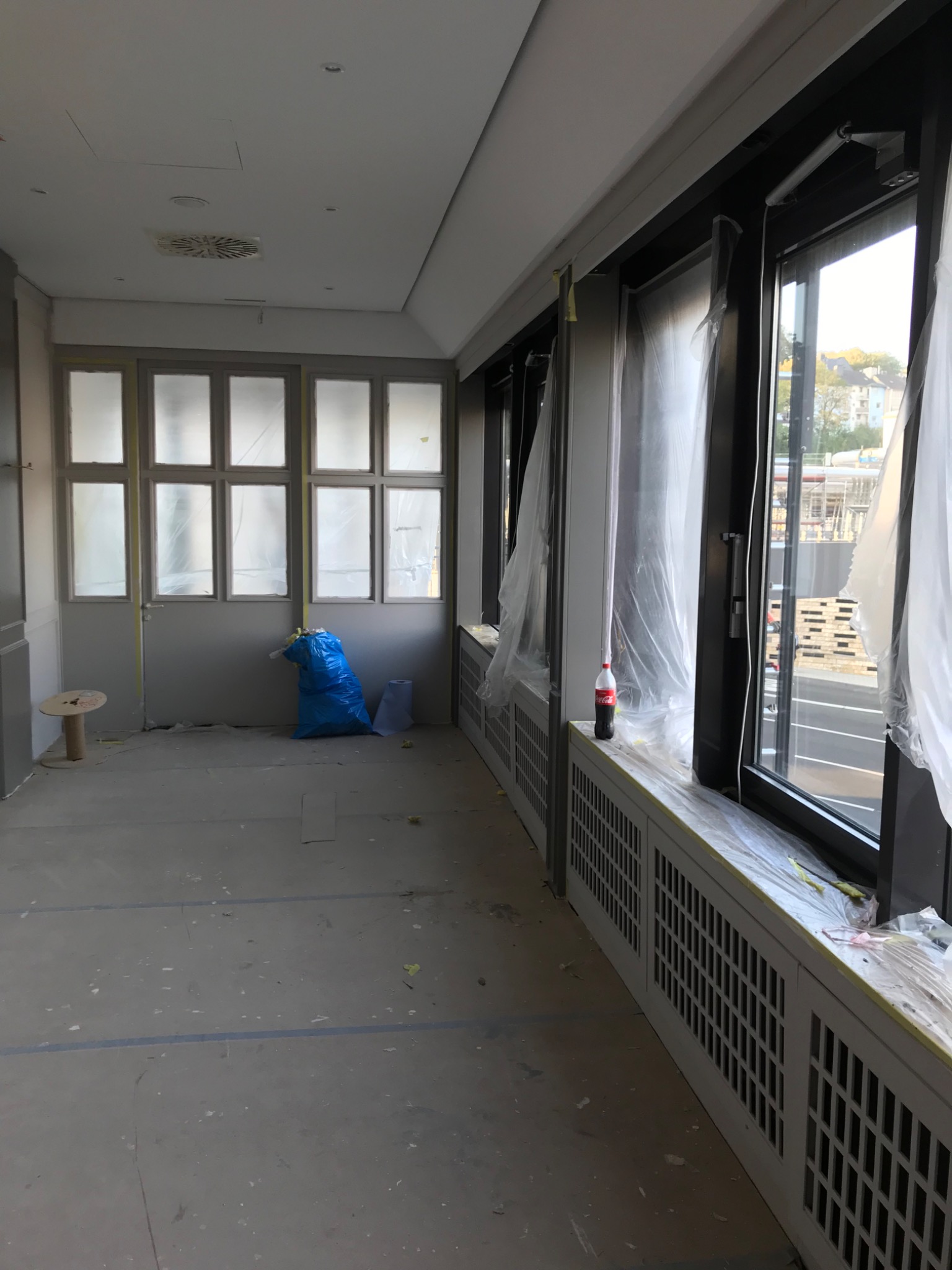 Przygotowania do prac wykończeniowych w pomieszczeniu w Warszawie: okna i grzejniki zabezpieczone folią malarską, drzwi oklejone taśmą, widoczny niebieski worek na odpady budowlane i butelka...