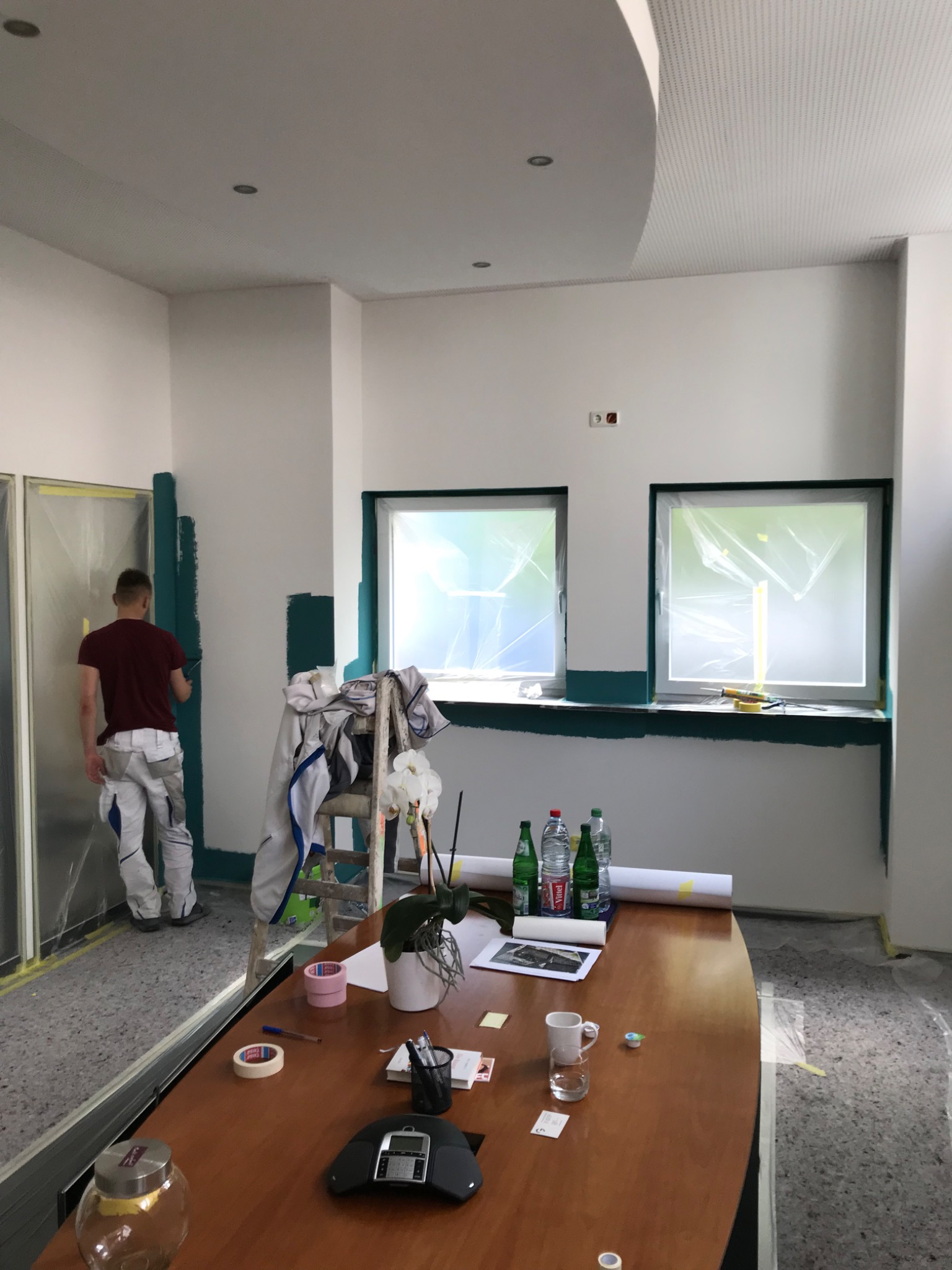 Mężczyzna w roboczym ubraniu maluje na zielono fragment ściany w biurze, zabezpieczone okna folią, na stole butelki wody, dokumenty i przybory biurowe.