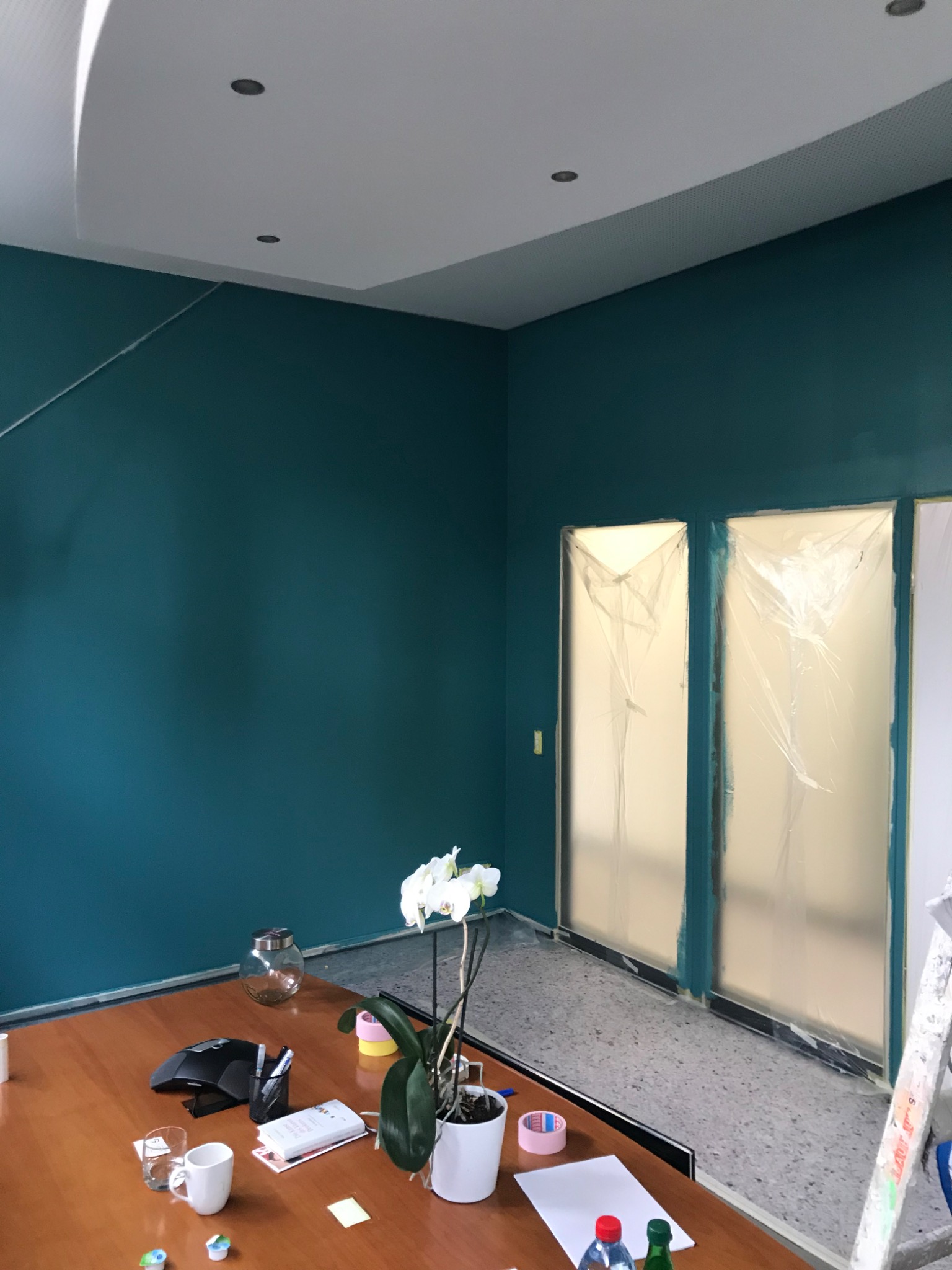 Pokój biurowy z pomalowanymi na turkusowo ścianami, zabezpieczonymi folią oknami, kwiatem orchidei na stole i widoczną drabiną, sugerujący trwające prace wykończeniowe.