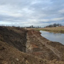 Brzeg rzeki z wykopanym korytem, ślady opon w błocie, pochmurne niebo.