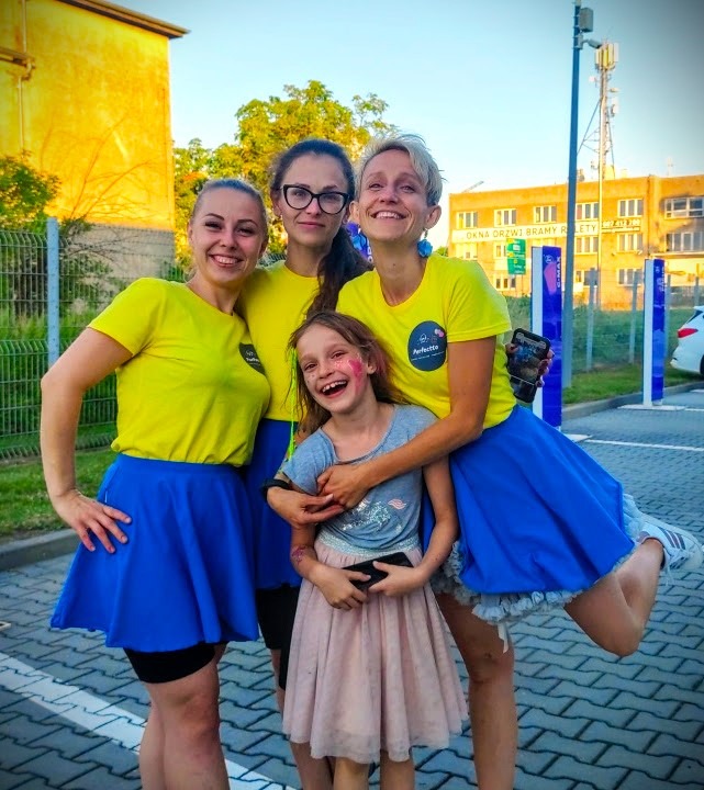 Grupa czterech kobiet i dziewczynka z pomalowaną twarzą, ubrane w żółte koszulki z logo i niebieskie spódniczki, pozuje do zdjęcia na parkingu przed budynkiem.