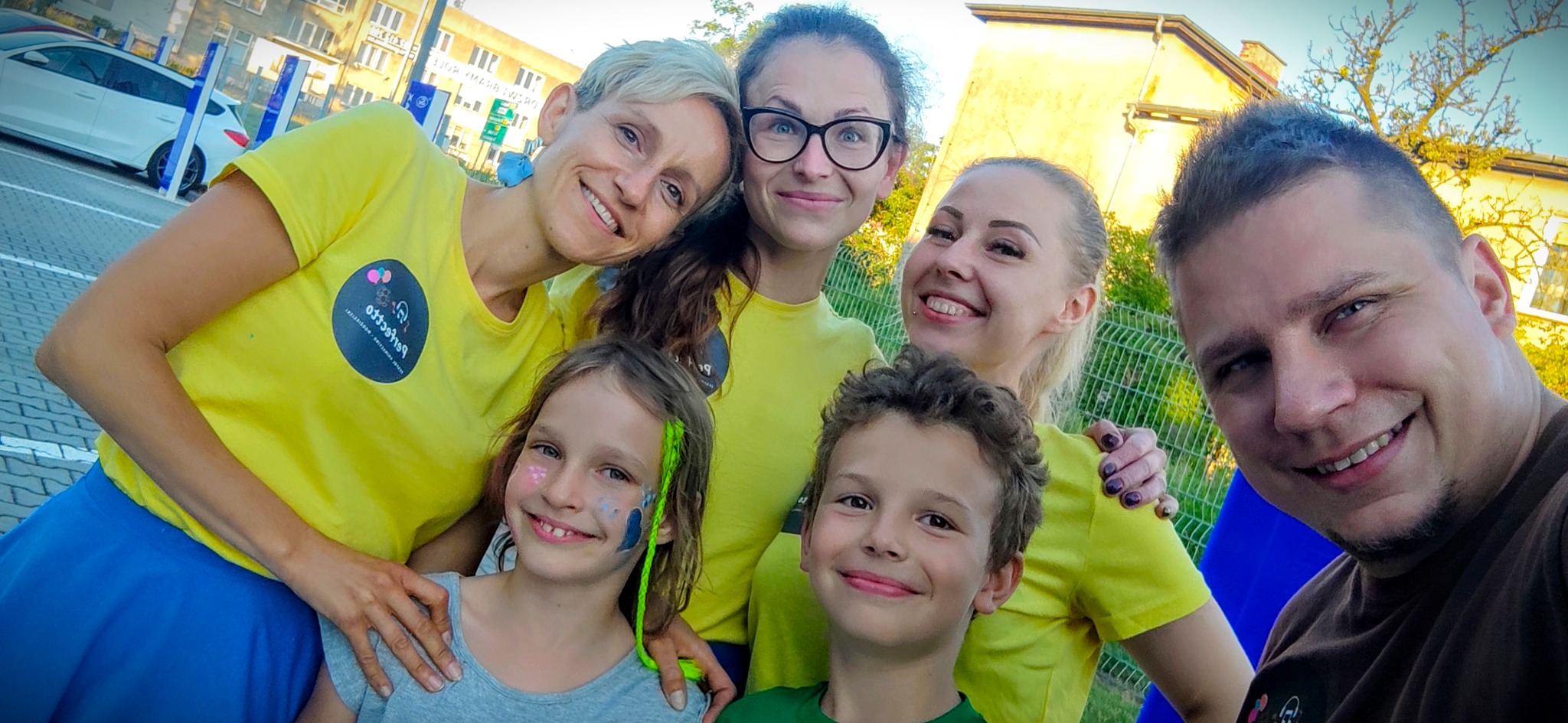 Grupa uśmiechniętych osób, w tym dwoje dzieci z pomalowanymi twarzami, pozuje do zdjęcia na zewnątrz, wszyscy mają na sobie żółte koszulki z logo firmy.