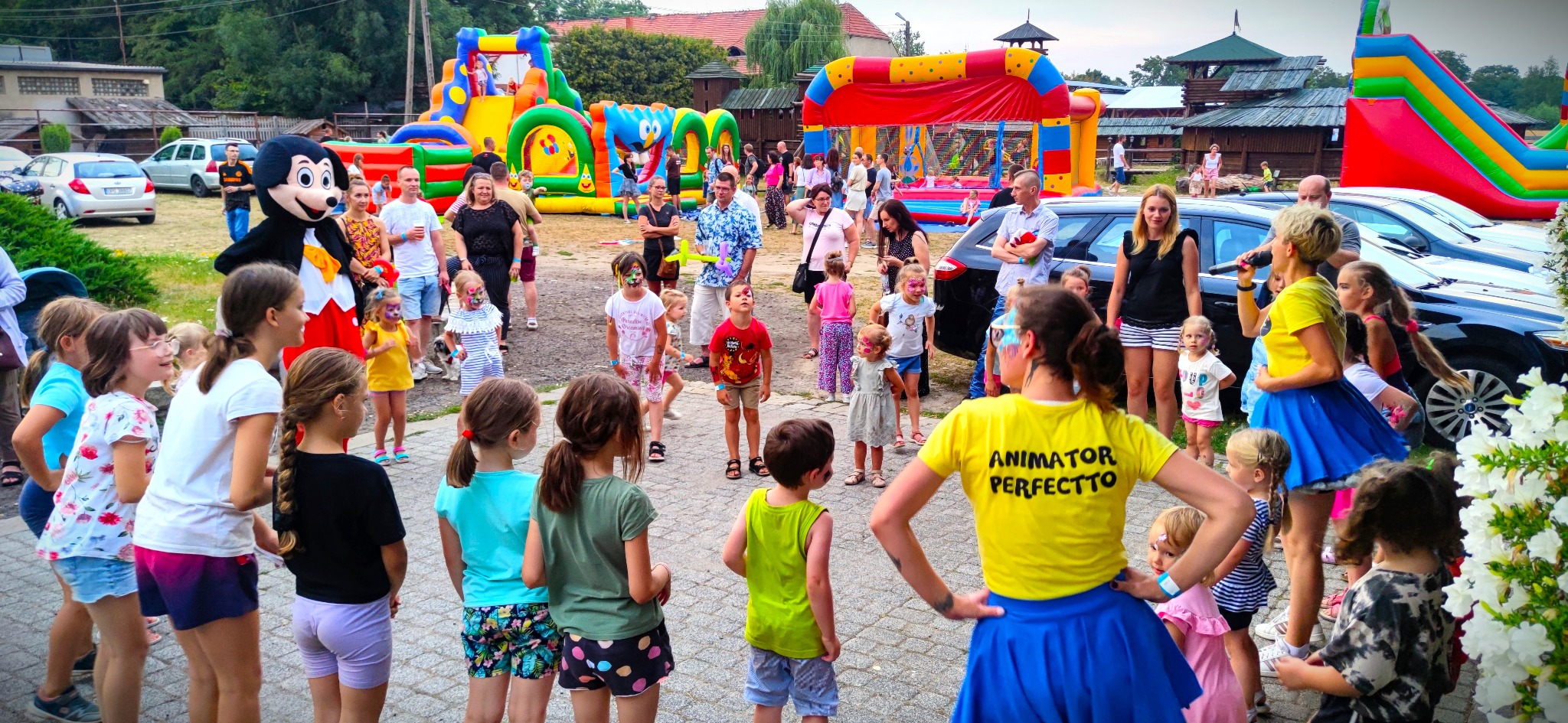 Grupa dzieci z pomalowanymi twarzami stoi przed dmuchanymi zamkami, animator w żółtej koszulce z napisem 'Animator Perfectto' prowadzi zabawę, w tle postać w kostiumie Myszki Miki i zaparkowane...