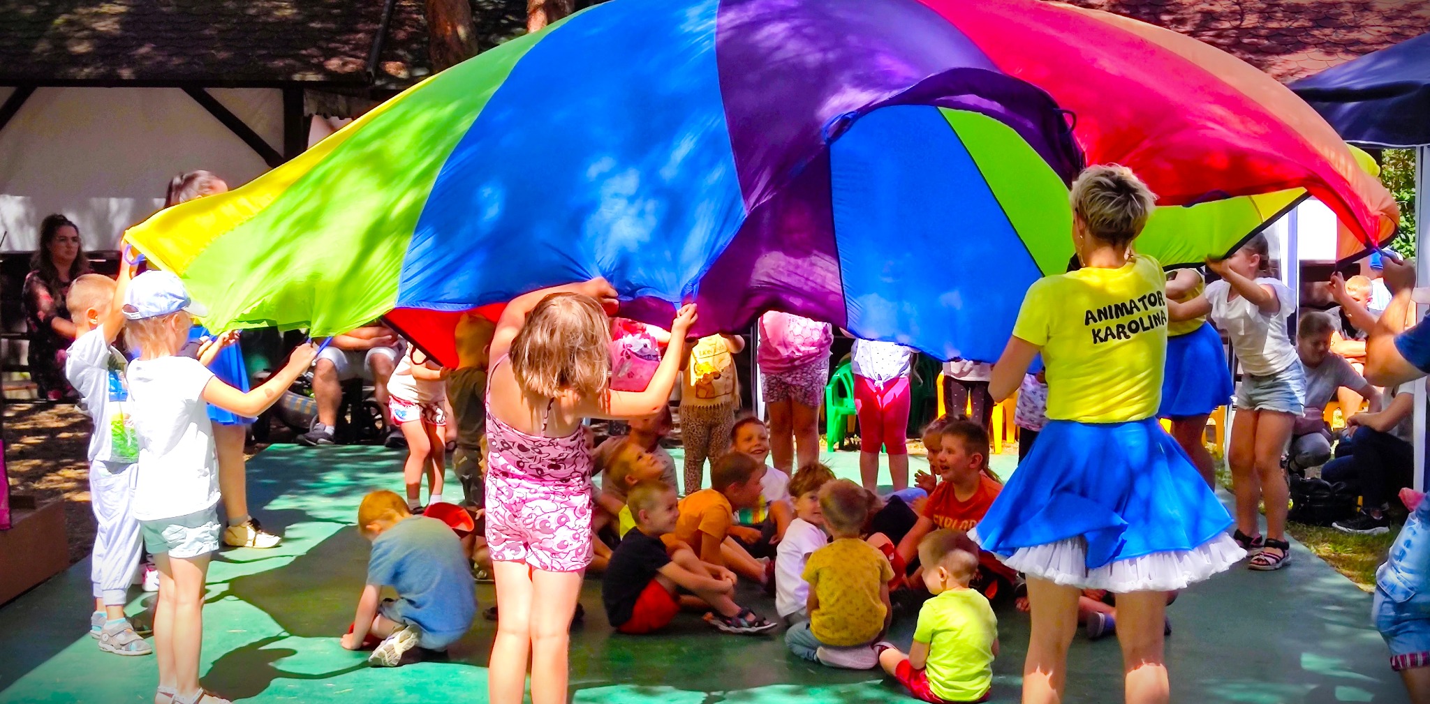 Grupa dzieci bawi się pod rozciągniętą, kolorową chustą animacyjną; widoczna animatorka w żółtej koszulce z napisem 'ANIMATOR KAROLINA'.