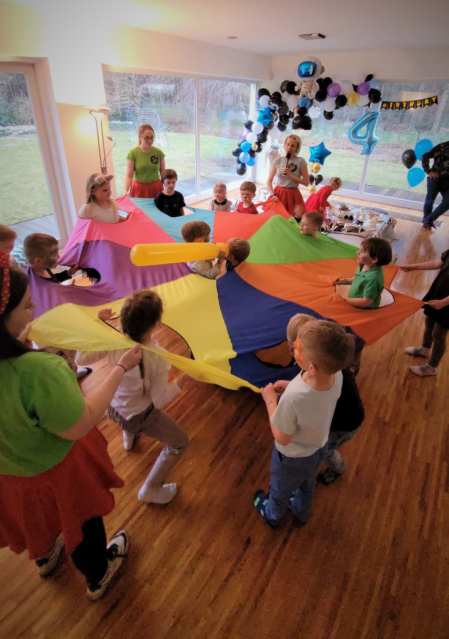 Grupa dzieci bawi się kolorową chustą animacyjną w pomieszczeniu udekorowanym balonami urodzinowymi, pod nadzorem dwóch animatorek w zielonych koszulkach i czerwonych spódnicach. Jeden z chłopców...
