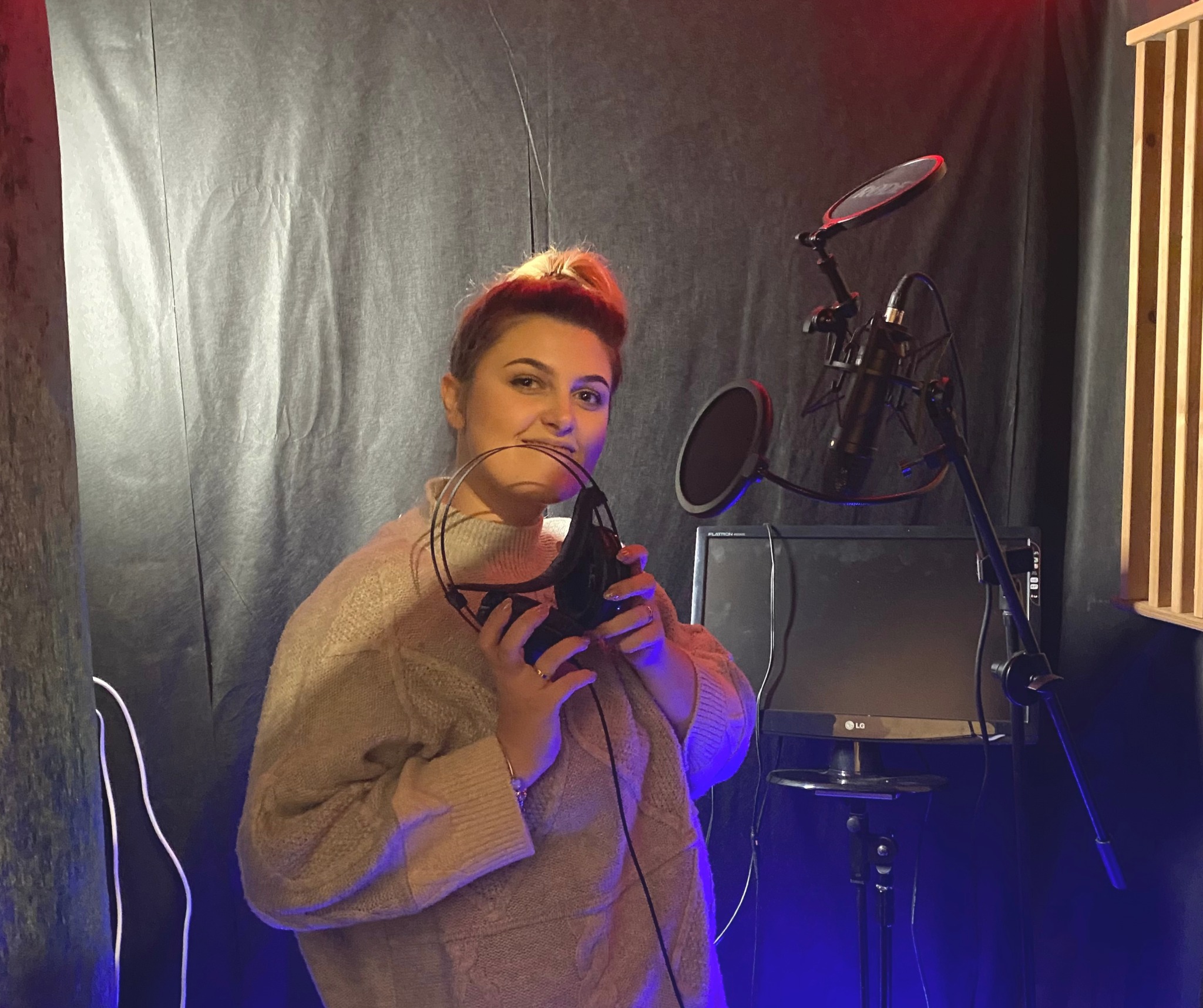 Kobieta trzyma słuchawki w dłoniach, w tle mikrofon z pop filtrem, monitor i panele akustyczne w amatorskim studio nagrań z czarnym tłem.