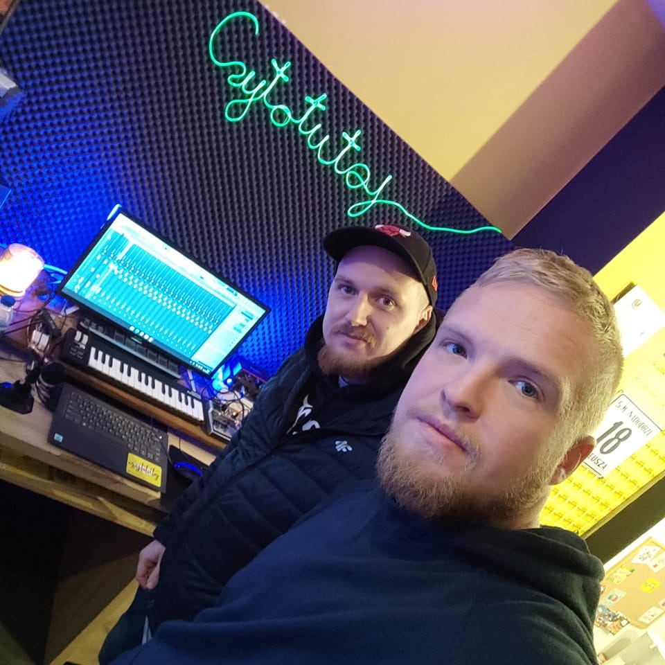 Dwóch mężczyzn w studiu nagrań, widoczny monitor z programem do obróbki dźwięku, klawiatura MIDI, ściany wygłuszone akustycznie, neonowy napis 'Cytotutoy' na ścianie, ujęcie z bliska.