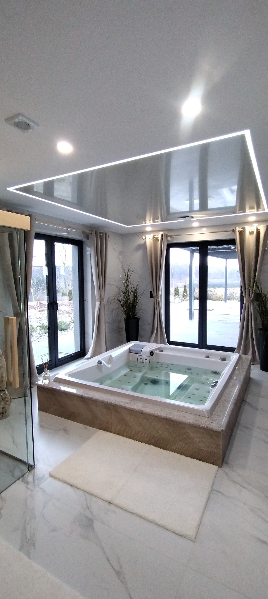 Luksusowa łazienka z wanną jacuzzi na podwyższeniu, marmurowa podłoga i ściany, podwieszany sufit z oświetleniem LED, duże okna z zasłonami, rośliny w donicach.