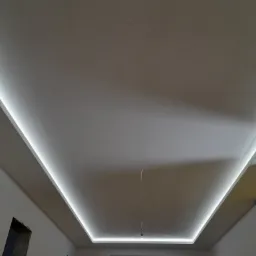 Sufit podwieszany z oświetleniem LED wzdłuż krawędzi, widoczne niedokończone wykończenia ścian i zabezpieczona folią część pomieszczenia.