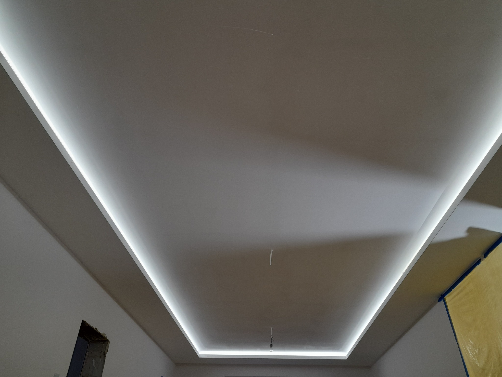 Sufit podwieszany z oświetleniem LED wzdłuż krawędzi, widoczne niedokończone wykończenia ścian i zabezpieczona folią część pomieszczenia.