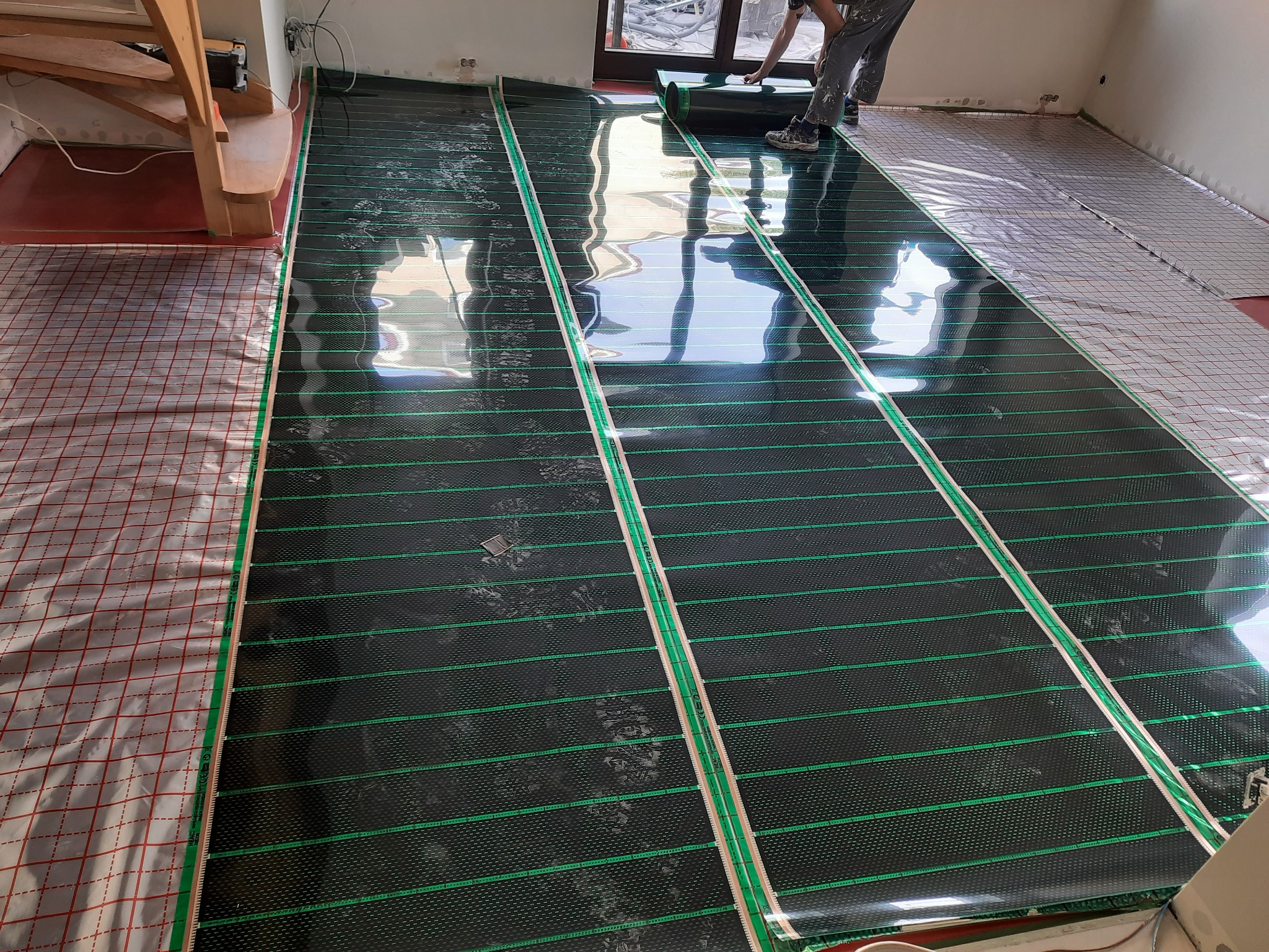 Instalacja mat grzewczych podłogowych na folii z siatką, widoczne fragmenty podłogi i schodów, osoba rozwija rolkę maty.