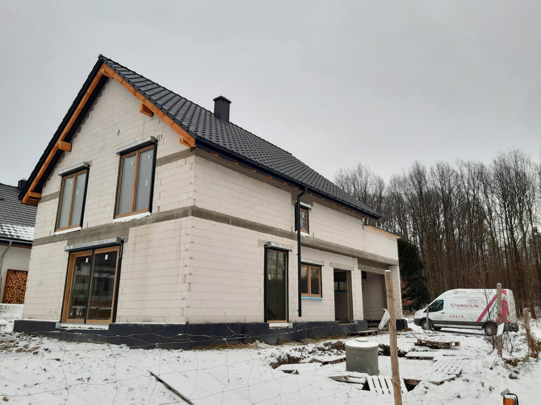 Surowy stan domu jednorodzinnego w zimowej scenerii, widoczne okna z brązowymi ramami, drzwi przesuwne z zamontowanymi roletami zewnętrznymi w kolorze antracytowym, biała ściana z betonu...