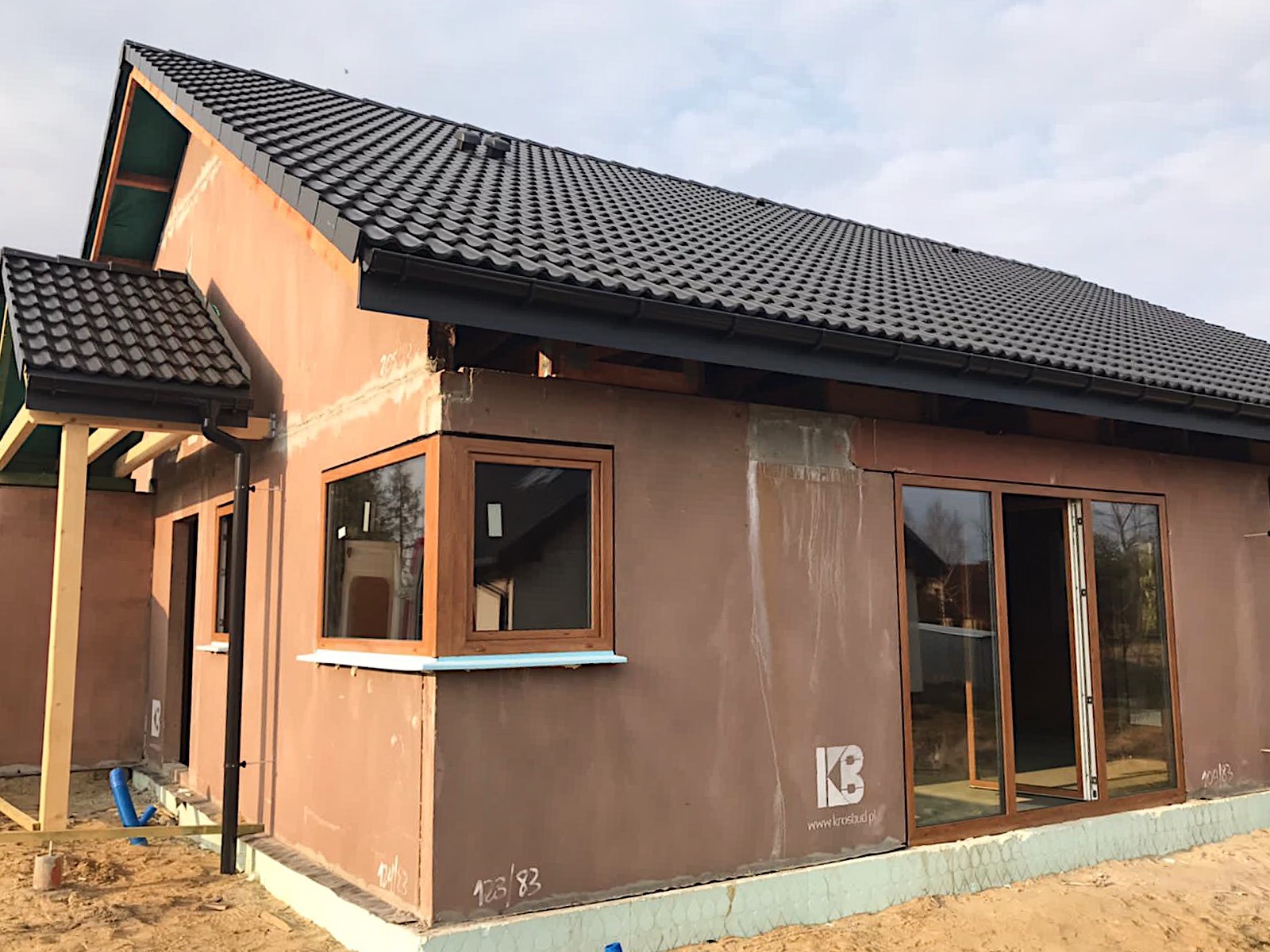 Dom w trakcie budowy z zamontowanymi oknami w drewnianych ramach i przesuwnymi drzwiami tarasowymi, widoczne ocieplenie budynku i czarna dachówka.