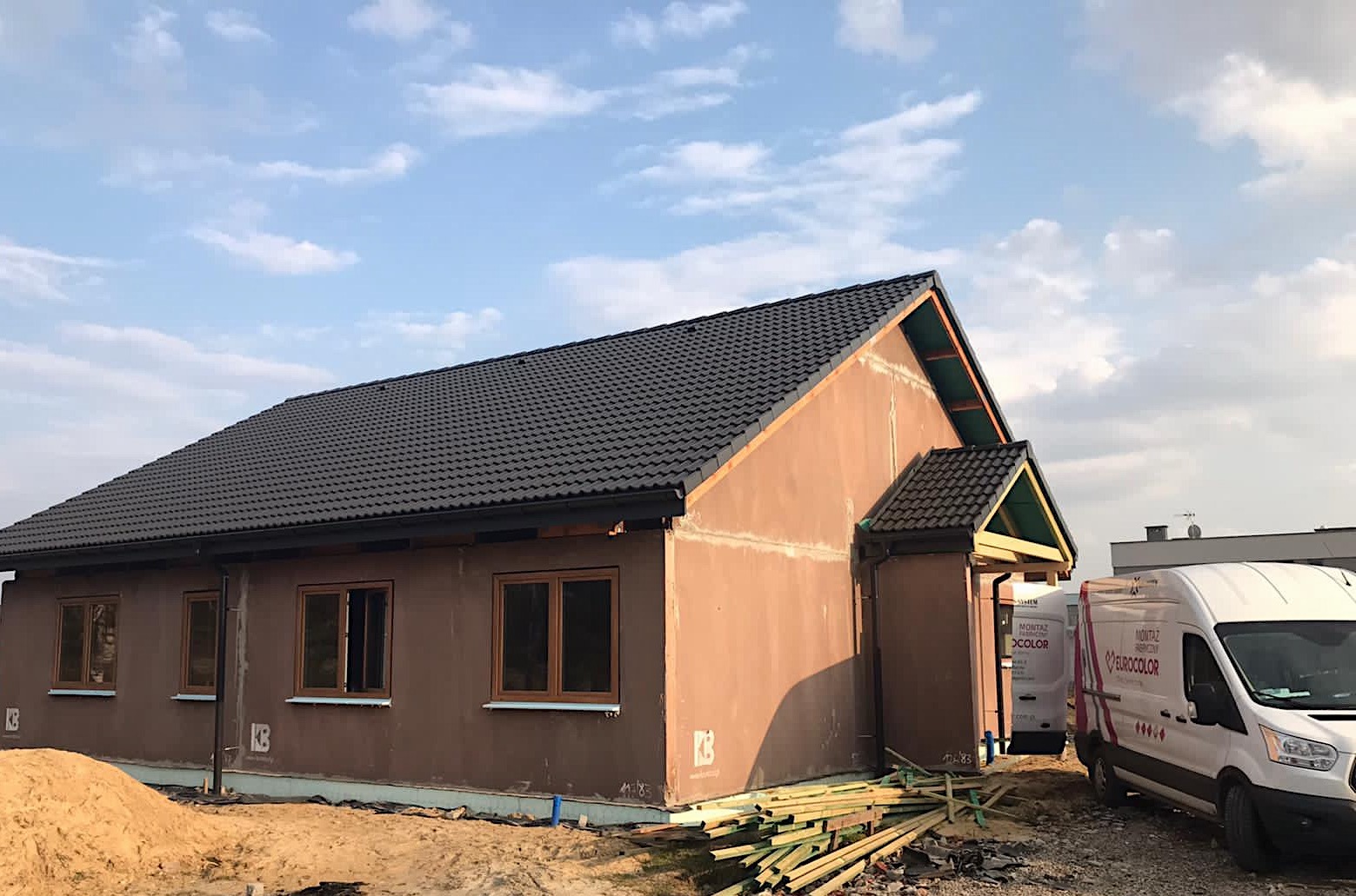 Nowo wybudowany dom jednorodzinny z ciemnym dachem, otynkowany na brązowo, z zamontowanymi oknami z brązowymi ramami, widoczne deski i samochody dostawcze w tle.