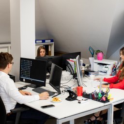 Biuro Rachunkowe TM PROGRES sp. z o.o. - Trzy osoby pracujące w biurze przy biurkach z komputerami; na biurkach widoczne kalkulatory, telefony, dziurkacz, przekąski na talerzyku i przybory biurowe.