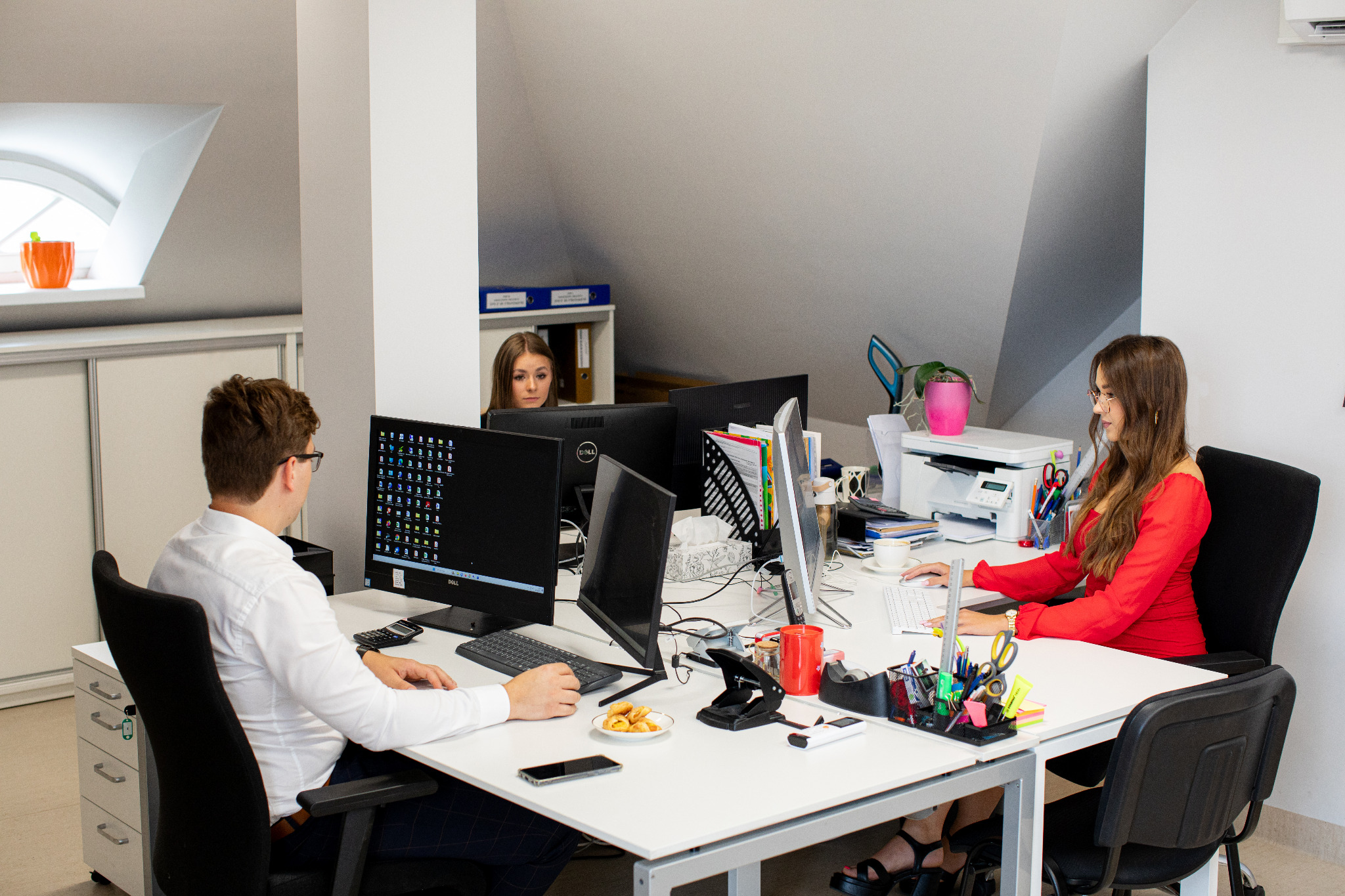 Trzy osoby pracujące w biurze przy biurkach z komputerami; na biurkach widoczne kalkulatory, telefony, dziurkacz, przekąski na talerzyku i przybory biurowe.