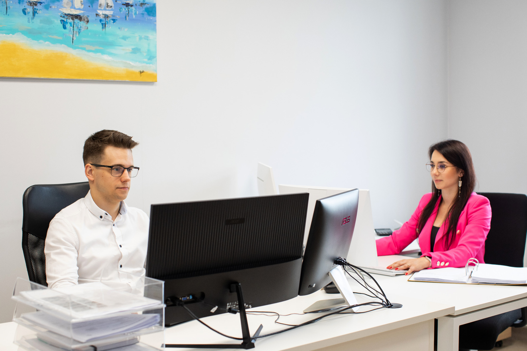 Dwie osoby pracują w biurze przy komputerach; na ścianie wisi abstrakcyjny obraz z motywem morza.