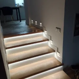 Schody drewniane z podświetleniem LED wbudowanym w stopnie i dodatkowym oświetleniem punktowym w ścianie, widok z dołu.