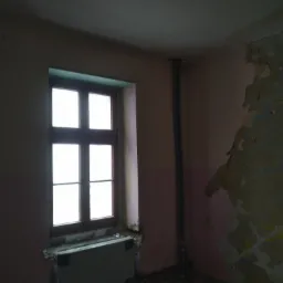 Wnętrze pokoju z oknem, ściany w trakcie demontażu, widoczne fragmenty starej farby i instalacje.