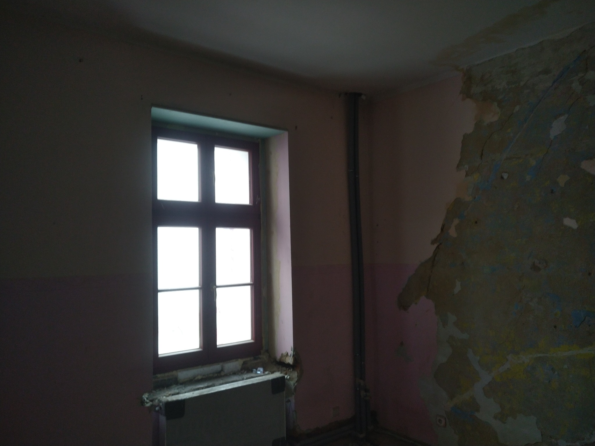 Wnętrze pokoju z oknem, ściany w trakcie demontażu, widoczne fragmenty starej farby i instalacje.