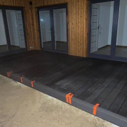 Green Building - Nowo wybudowany, ciemny, drewniany podest z pomarańczowymi taśmami zabezpieczającymi, przylegający do budynku z drewnianą elewacją i trzema parami przeszklonych drzwi.