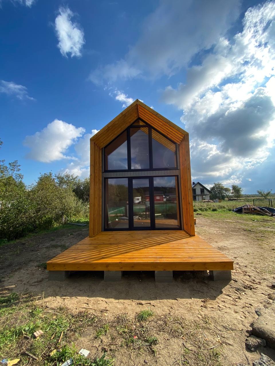Nowoczesny, drewniany domek letniskowy z dużymi oknami i tarasem, posadowiony na betonowych bloczkach, na tle błękitnego nieba z chmurami.