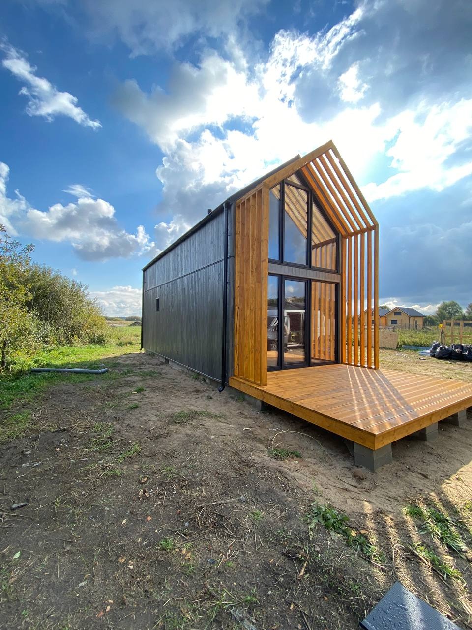 Nowoczesny dom modułowy z ciemną elewacją i drewnianą konstrukcją szkieletową oraz tarasem z desek, widok z poziomu gruntu, błękitne niebo z obłokami w tle.