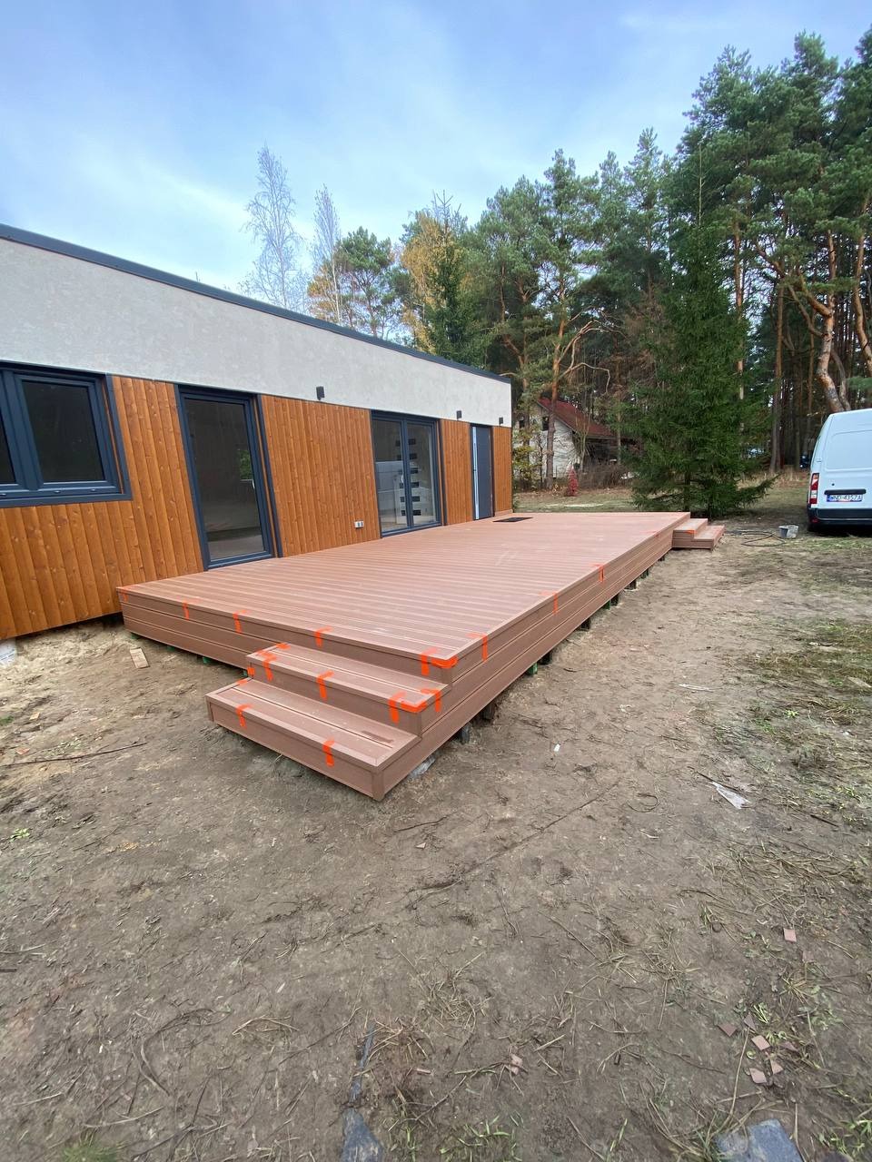 Nowo wybudowany, duży, brązowy taras z kompozytu z trzema schodkami, zabezpieczony pomarańczową taśmą, przylegający do nowoczesnego domu z drewnianą elewacją, na tle lasu.