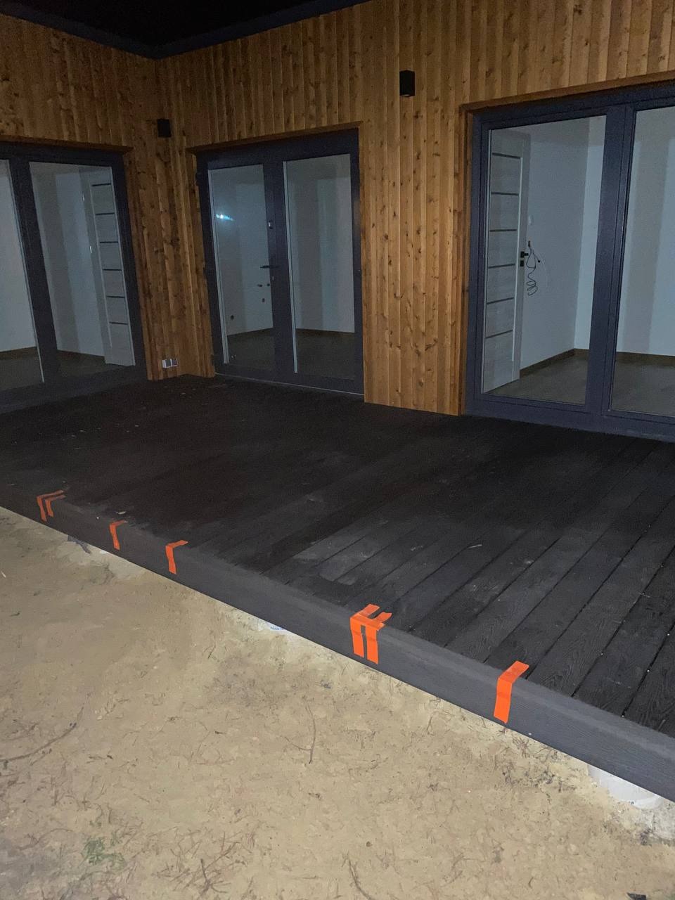 Nowo wybudowany, ciemny, drewniany podest z pomarańczowymi taśmami zabezpieczającymi, przylegający do budynku z drewnianą elewacją i trzema parami przeszklonych drzwi.