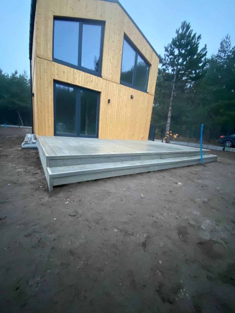 Nowoczesny dom z drewnianą elewacją i szarym tarasem z dwoma stopniami, na jeszcze niezagospodarowanym terenie.