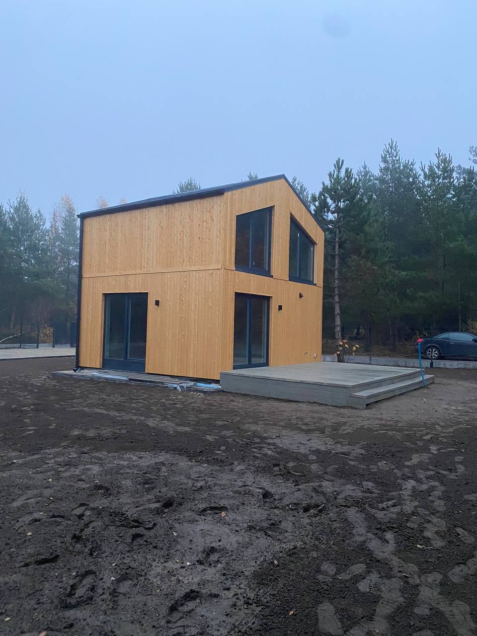 Nowoczesny, dwukondygnacyjny dom z elewacją z jasnego drewna i czarnymi ramami okiennymi, z przylegającym tarasem z kompozytu w kolorze szarym, otoczony ziemią i drzewami.